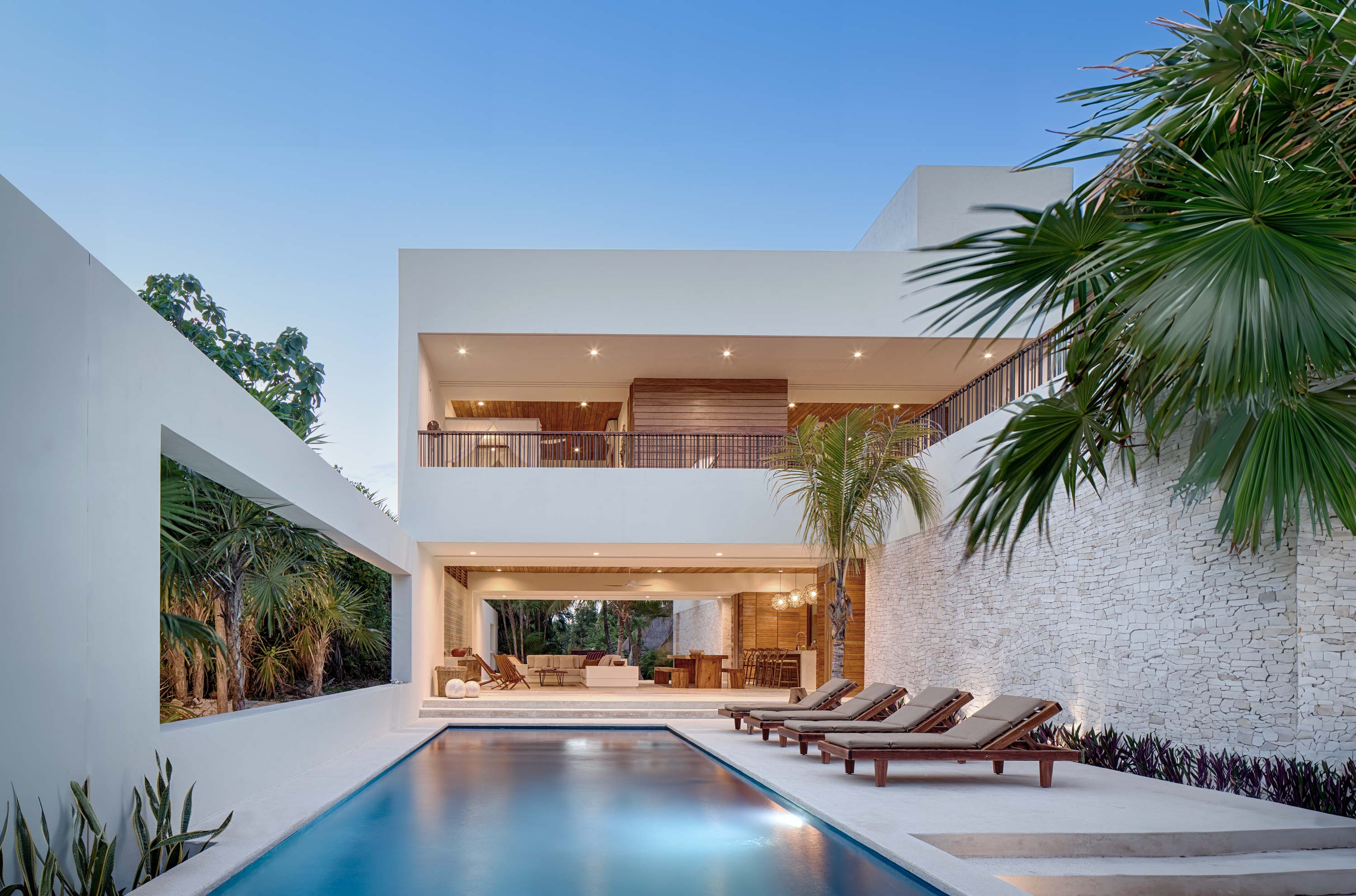 Exterior photo of Casa Xixim by Specht Novak Architects. Shot by Taggart Sorensen, featuring the pool, lounging area, and views of the interior and upper level of the property framed by palm trees.