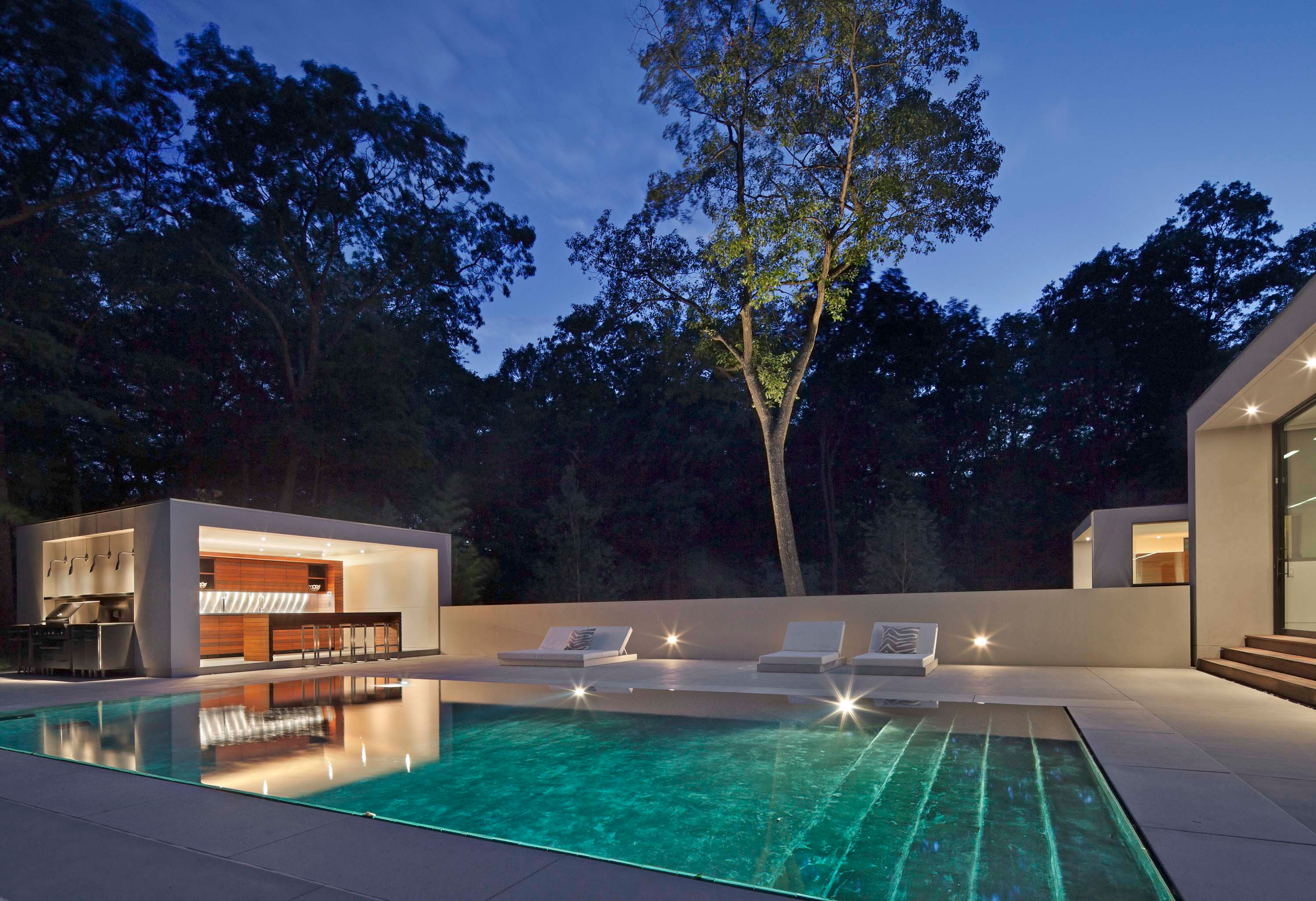 Exterior photo of the New Canaan Residence by Specht Novak Architects. Shot at dusk by Elizabeth Felicella featuring the pool, lounging area, and covered outside dining/bar area surrounded by trees.