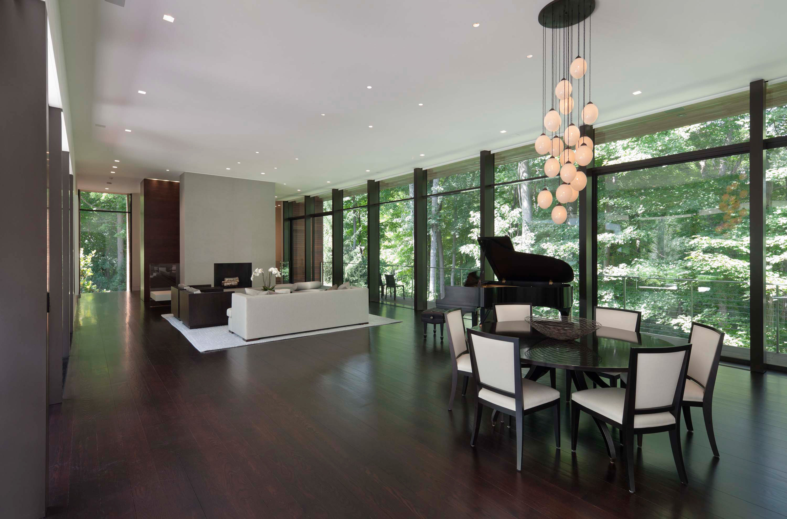 Interior photo of the New Canaan Residence by Specht Novak Architects. Shot by Elizabeth Felicella featuring an open living space, lavish chandelier, piano, and dining area surrounded by glass walls that welcome the surrounding nature to the space.
