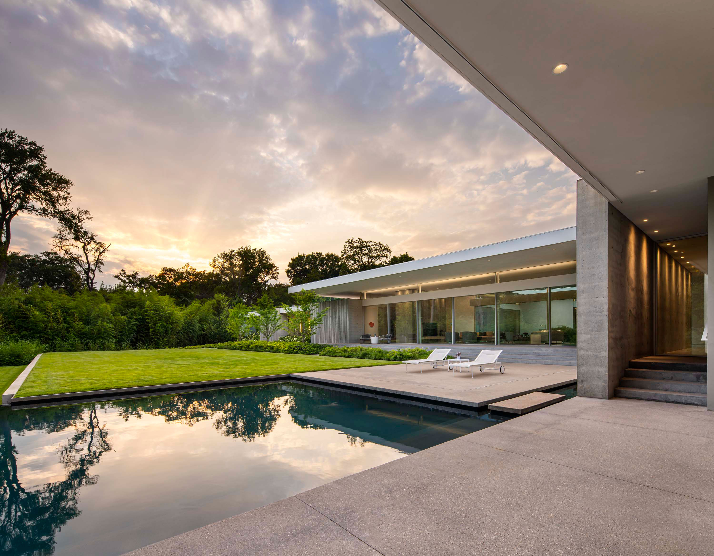 Exterior photo of the Preston Hollow Residence by Specht Novak Architects. Shot at sunrise by Manolo Langis, featuring an entry hallway, the pool, yard, lounging areas, and glowing home.